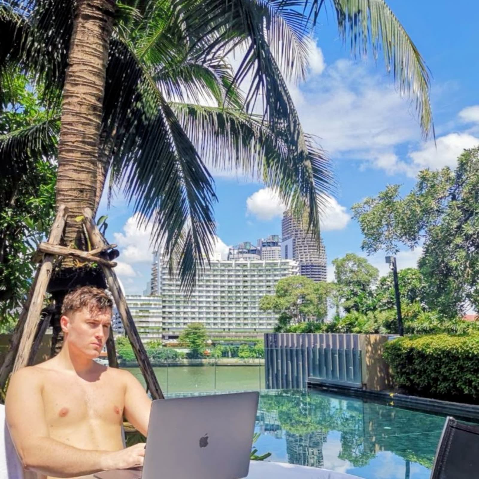 shirtless man working on a laptop poolside under a palm tree, city skyline behind