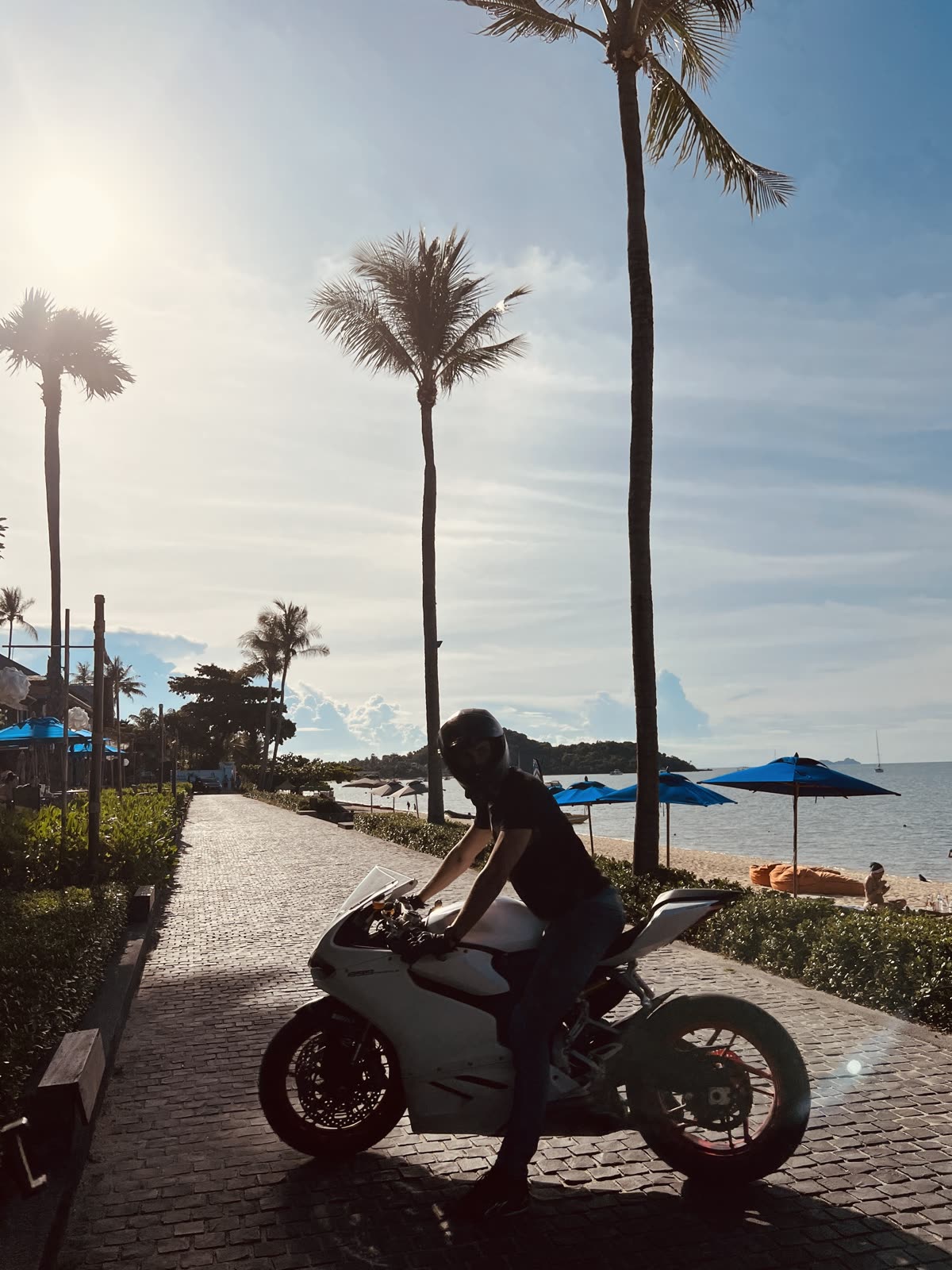 man standing beside a white Ducati motorcycle on a beachfront promenade with palm trees