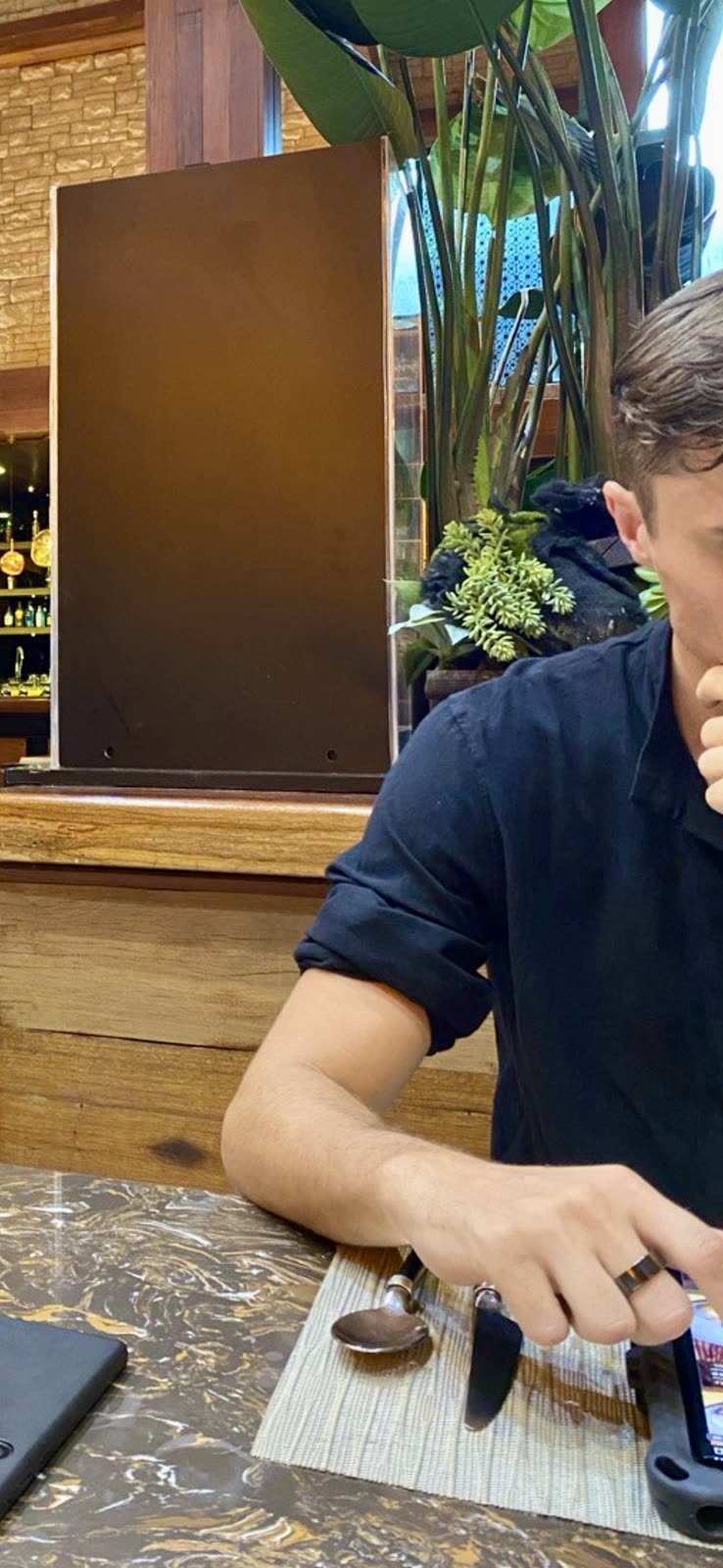 man seated at a restaurant table using his phone, plants and dark wood decor around him